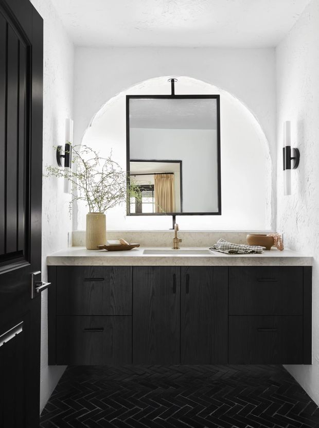 A compact, refined bathroom vanity nook with a wall-mounted dark timber cabinet, pale stone countertop, integrated basin and brass tapware. The space features white textured plaster walls, a large black-framed rectangular mirror set within an arched wall recess, black wall sconces, and a dark herringbone tile floor. A black paneled door is visible at left.
