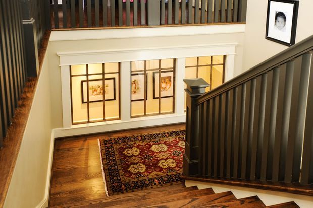 Interior view from a staircase landing looking down toward a hallway or entry area. The space features dark stained timber stair treads and balustrades, warm timber flooring, cream painted walls, decorative white window trim, and large multi-pane windows with framed artwork. A richly patterned oriental rug adds colour and texture. The staircase and upper landing dominate the composition.