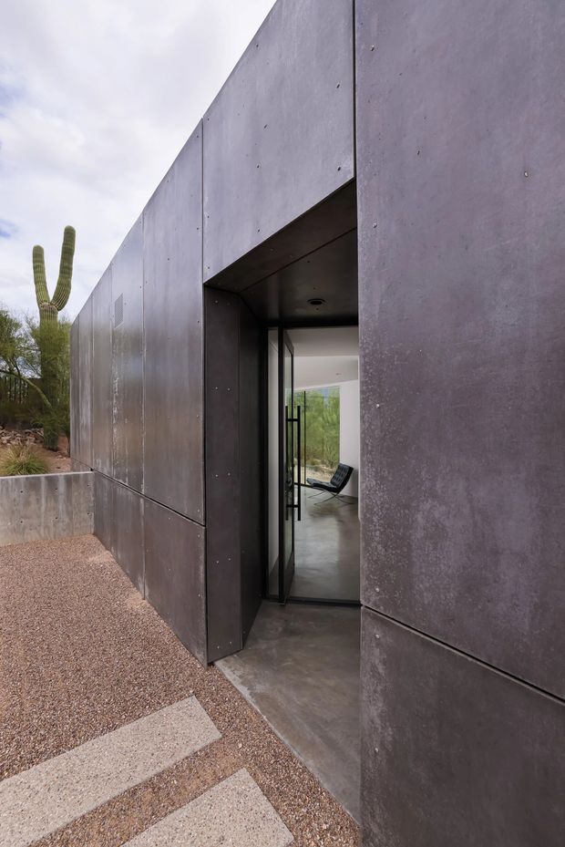 Exterior view of a minimalist, monolithic building clad in dark grey metal panels, with a recessed glazed entry and a simple concrete threshold. The building sits in a gravelled desert-style landscape with a sculptural cactus visible beside it. Through the open glass door, a polished concrete interior and black lounge chair can be seen.