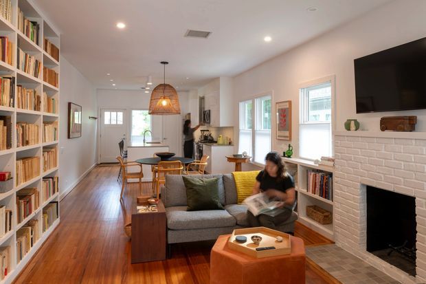 Warm, open-plan residential interior combining a living room, dining area, and kitchen. The room features extensive built-in white bookshelves, polished timber flooring, a grey sofa with colourful cushions, a tan leather ottoman, a white-painted brick fireplace with a wall-mounted TV, and a woven pendant light over the dining table. Large windows provide natural light and the space has a welcoming, lived-in character.