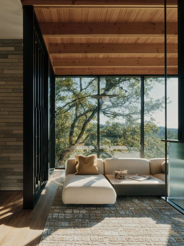 A warm, modern living room with a low-profile cream sectional sofa positioned beside expansive black-framed glazing. The room features a richly timber-lined ceiling with exposed beams, dark horizontal brick wall cladding, timber flooring, and a large muted patterned rug. Floor-to-ceiling windows overlook mature trees and a distant landscape, while sunlight creates strong shadows across the seating area and rug. A slim pendant light and dark metal framing add architectural definition.
