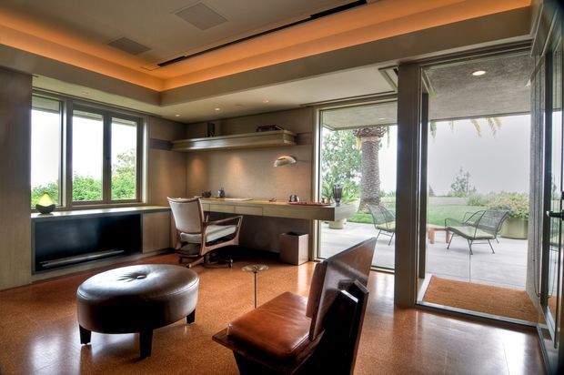 A refined open-plan home office and lounge with a built-in wood desk, task chair, low dark cabinetry, leather lounge chair, and round upholstered ottoman. Large windows and fully glazed sliding doors open onto a furnished outdoor terrace. The room features warm recessed cove lighting, timber-toned surfaces, and polished brown flooring.