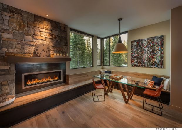 Warm, modern dining and living area featuring a dramatic stacked-stone fireplace wall with a linear firebox, timber flooring, and a built-in window-side dining nook. A glass-topped timber dining table with contemporary leather chairs sits beneath a large pendant light, while expansive corner glazing overlooks trees. Abstract artwork and a sculptural clock add visual interest.