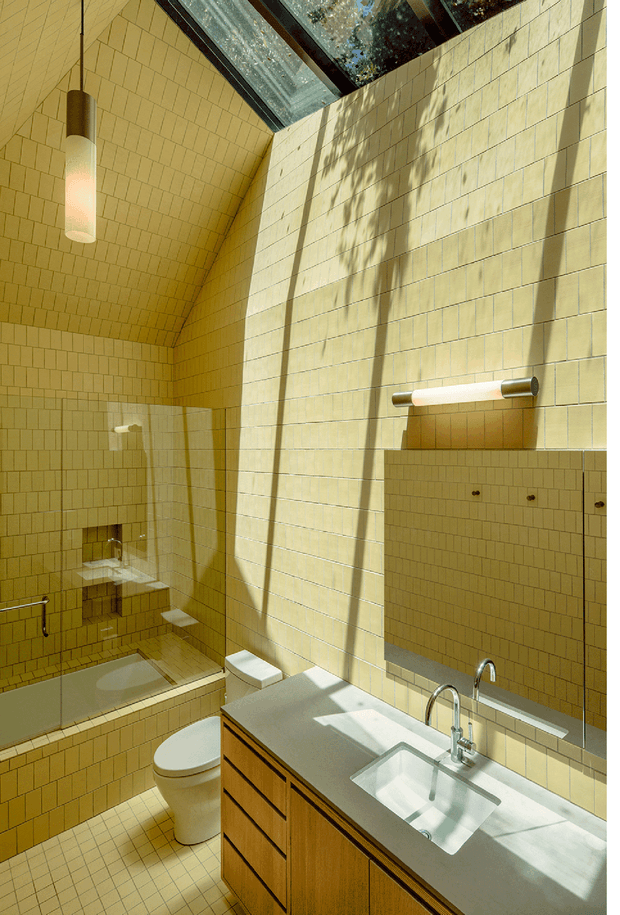 A sunlit contemporary bathroom with warm yellow-beige rectangular wall and floor tiles, a tiled bathtub and shower enclosure, white toilet, and a timber vanity with a pale solid benchtop and integrated white basin. A large mirror, wall-mounted linear light, cylindrical pendant, and skylight create strong natural illumination and shadows.