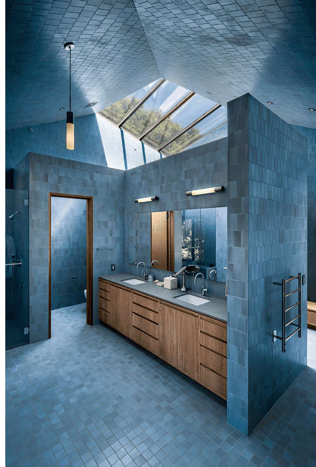 A spacious contemporary bathroom clad in blue-grey square tiles from floor to ceiling, with a large skylight bringing in natural light. The room features a long double vanity with warm timber cabinetry, grey benchtop, multiple basins and wall-mounted fixtures, a large mirror, open shower area, towel ladder, and a strong blue-toned palette.
