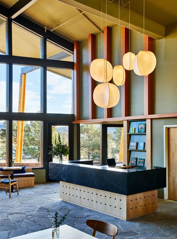 A dramatic, double-height reception or lobby with expansive black-framed glazing, a sculptural dark stone reception desk, built-in timber and cream cabinetry, blue open shelving, and a cluster of rounded pendant lights. Vertical timber-toned wall fins add warmth against muted green walls, while irregular natural stone flooring and views of trees create an earthy, refined atmosphere.