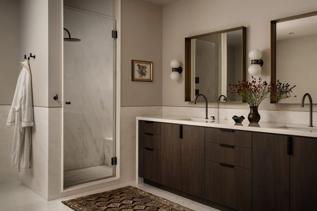 A refined contemporary bathroom with a double vanity, dark wood cabinetry, pale stone benchtop, twin wall mirrors, black tapware and sconces, and a glass-enclosed marble-look shower. Neutral beige walls and warm, subdued lighting create a calm, luxurious atmosphere.