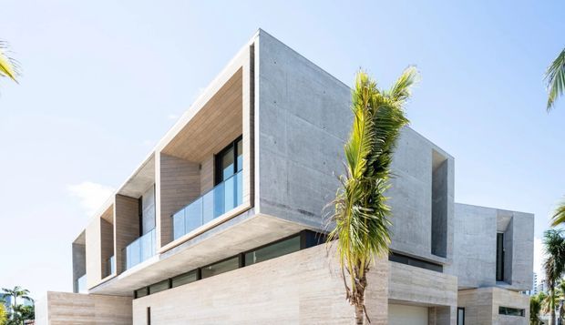 A contemporary multi-level concrete residence viewed from outside in bright daylight. The angular facade combines smooth board-formed or rendered concrete walls and projecting slabs with warm timber soffits, large dark-framed glazed openings, and glass balcony balustrades. Palm trees are visible around the building, giving the setting a sunny, tropical character.
