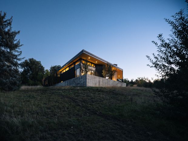 A modern single-storey house viewed from a grassy, wooded site at dusk. The building has a strong angular roof plane, extensive warm-lit glazing, dark exterior wall surfaces, and a raised concrete retaining or boundary wall. Trees and vegetation surround the house, creating a secluded setting.