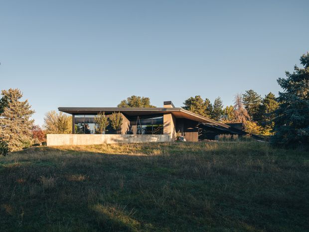 A low, single-storey modern house set within a grassy, wooded landscape. The building has broad horizontal proportions, extensive dark-framed glazing, warm timber-toned wall surfaces, and a dramatically projecting shallow roof with deep eaves. Viewed from a distance across the sloping lawn in warm late-day light.