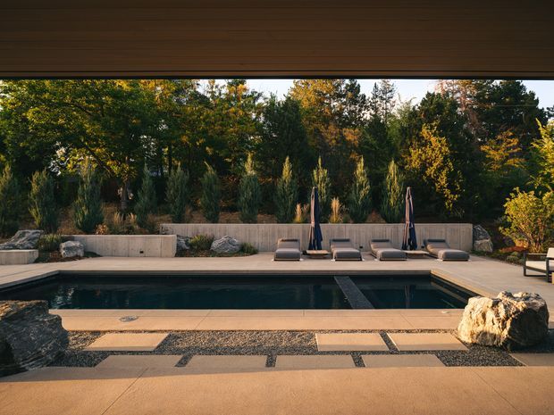 A tranquil outdoor swimming pool viewed from beneath a broad timber-lined roof overhang. The rectangular pool is surrounded by warm-toned concrete paving, dark gravel bands, stone stepping slabs, large natural boulders, and a minimalist lounge area with sun loungers and closed umbrellas. Dense landscaped planting forms a private green backdrop.