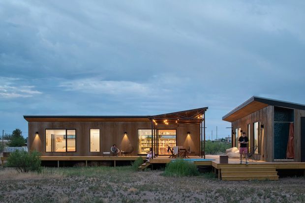 Exterior view of a contemporary modular or prefabricated home composed of connected timber-clad volumes. The low-profile dwelling sits on a raised timber deck in a sparse landscape, with broad glazed openings, a covered outdoor dining area illuminated by string lights, and a sloping roof on the right-hand volume. The scene is photographed at dusk under a cloudy blue sky.