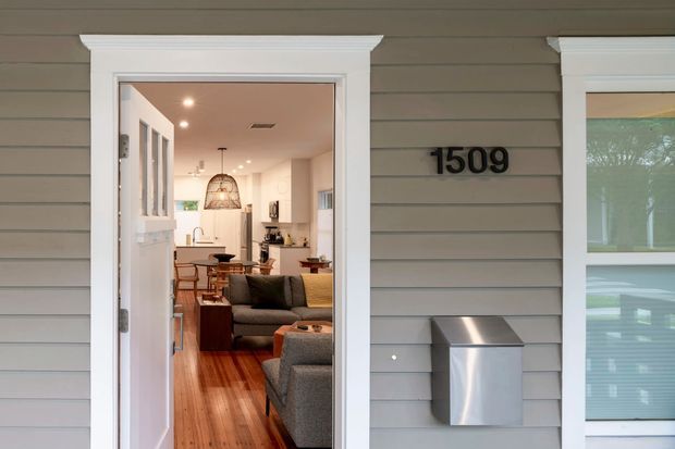 Front exterior view of a compact residential home with grey horizontal weatherboard cladding, white-trimmed entry door and window, and a modern metal wall-mounted mailbox. The open doorway reveals a warm, narrow interior with timber flooring, a living area, and a kitchen/dining space beyond.