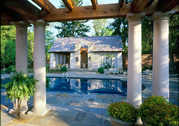 A landscaped outdoor pool courtyard viewed through a timber pergola with large classical columns. The rectangular swimming pool is surrounded by slate-like stone paving, clipped shrubs, potted plants and outdoor seating. Beyond the pool is a small traditional pavilion or pool house with a pitched, weathered roof, pale stucco walls and a central entry. Mature trees surround the setting, creating a secluded garden atmosphere.
