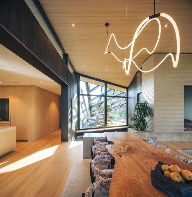 A spacious contemporary dining area with a large live-edge timber dining table and patterned upholstered chairs. The room features warm timber flooring and ceiling boards, dark textured wall cladding, pale brickwork, extensive black-framed glazing, and a sculptural suspended pendant light. An open kitchen or living area is visible to the left, with landscaped greenery seen through the windows.