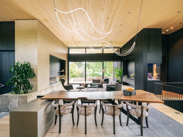 A sophisticated open-plan living and dining area with a long timber dining table and upholstered dining chairs in the foreground, flowing into a lounge with a sofa and armchairs. The room features a warm timber-lined ceiling, dark vertical wall panelling and cabinetry, pale stone or plaster finishes, large floor-to-ceiling windows, and sculptural suspended white lighting. A stair opening and dark built-in joinery are visible on the right, with greenery outside the glazing.