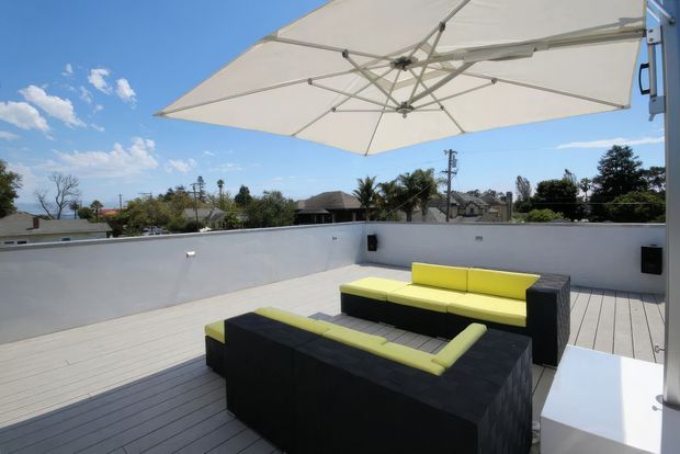 A bright rooftop terrace or deck furnished with black outdoor lounge seating accented by vivid lime-green cushions. A large white cantilevered umbrella provides shade, while white parapet walls, pale decking, built-in speakers, and a sunny suburban skyline create a relaxed outdoor entertaining setting.