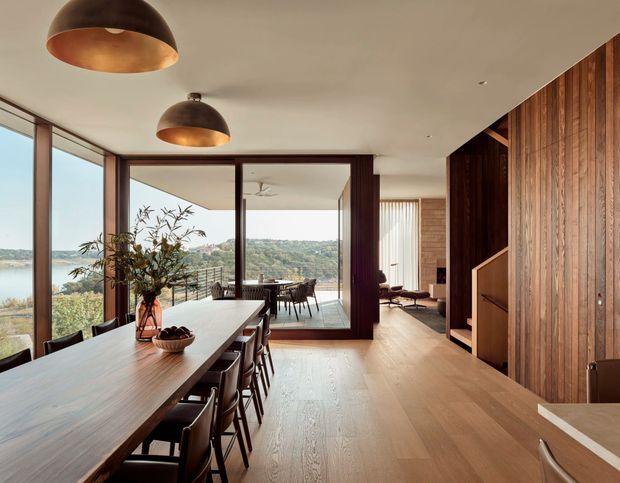 Warm contemporary open-plan dining and living area with a long timber dining table, dark dining chairs, exposed timber flooring, extensive floor-to-ceiling glazing, and a full-height timber feature wall with integrated joinery. The room opens toward a glazed terrace or balcony with a leafy scenic outlook. Large bronze pendant lights add a refined accent.