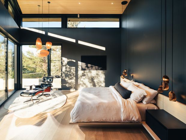 A contemporary bedroom with a low platform bed, dark vertically panelled walls, warm timber ceiling, extensive floor-to-ceiling glazing, and a compact sitting area with a black leather lounge chair and ottoman. Strong natural sunlight creates graphic shadows across the pale timber floor and walls. Integrated bedside joinery and sculptural pendant lights add a refined, modern feel.