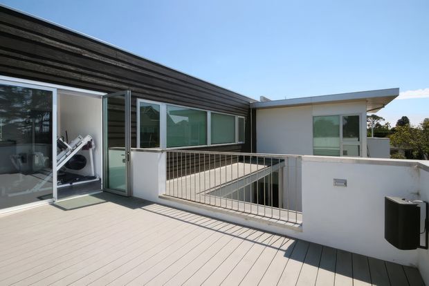 Bright contemporary rooftop terrace or upper-level outdoor deck beside a modern flat-roof building. The terrace has pale timber-look decking, white rendered parapet walls, a simple metal balustrade, large sliding glazed doors, and dark horizontal timber cladding. An exercise room or gym equipment is visible through the glazing, with a secondary white-clad volume and flat projecting roof beyond.