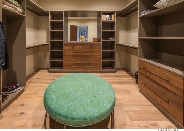 Walk-in wardrobe with extensive built-in open shelving, hanging spaces, and timber drawer cabinetry arranged around the room. A central round upholstered ottoman sits on light timber flooring, with warm recessed lighting and a mirrored central cabinet area.