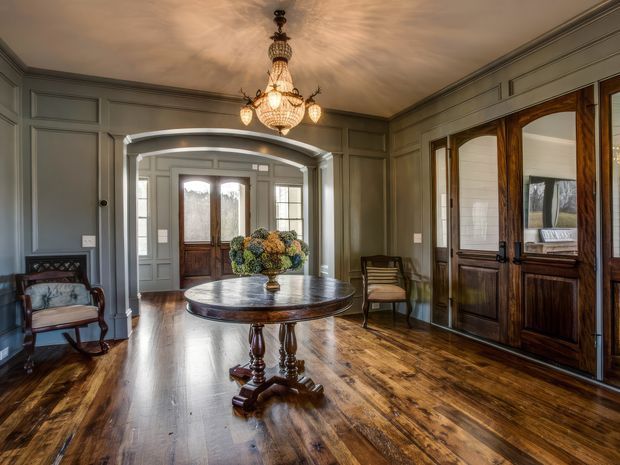 Elegant traditional entry hall or reception space with grey decorative wall panelling, a polished dark timber floor, ornate double entry doors with sidelights, large dark timber glazed doors, and a prominent crystal chandelier. A round pedestal table with floral arrangement anchors the room, with occasional upholstered seating along the walls. The space has a formal, historic character and warm natural light.