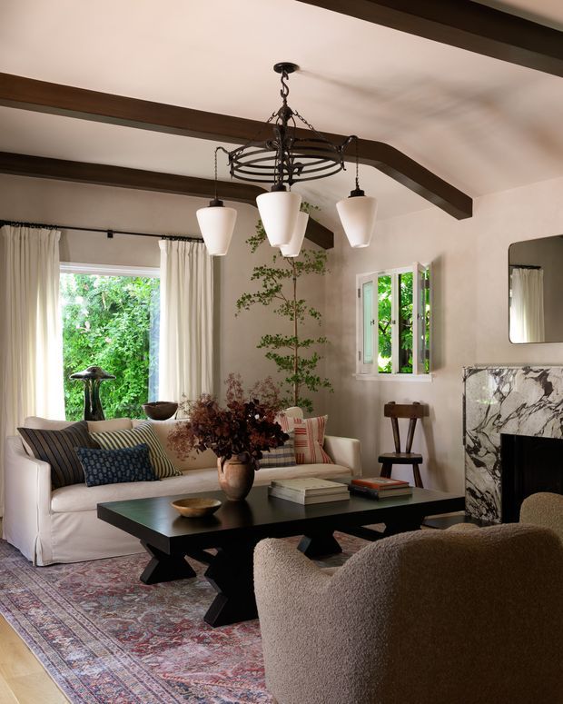 A warmly styled living room with exposed dark timber ceiling beams, cream plaster walls, a large patterned area rug, slipcovered sofa, sculptural lounge chairs, and a substantial dark wood coffee table. A marble-clad fireplace with dark veining anchors the right side of the room, while an ornate suspended metal chandelier and matching pendant lights provide lighting. Large windows with sheer curtains overlook lush greenery.