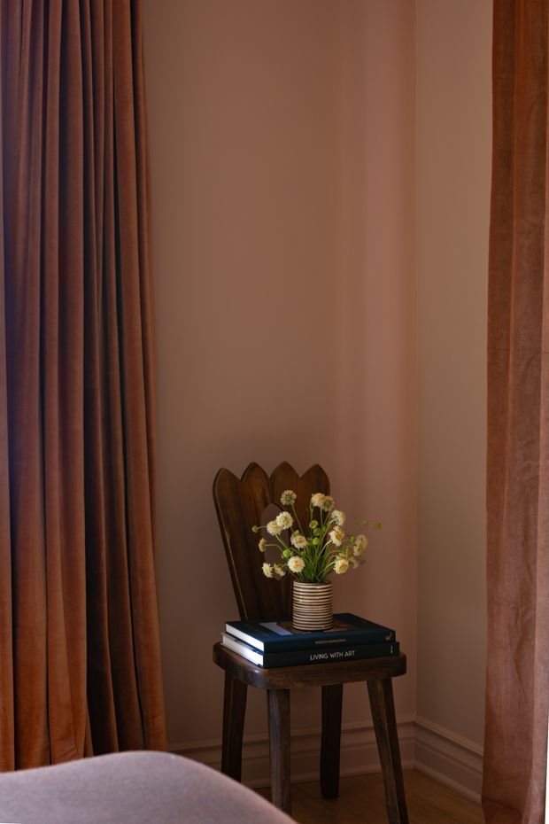 A warmly lit interior vignette showing a small dark wood side table with turned legs, stacked art books, a striped ceramic vase of dried flowers, and a carved wooden decorative object. The setting has muted blush-brown walls, dark timber flooring, floor-length rust and mauve curtains, white skirting boards, and part of a softly upholstered seating piece in the foreground.