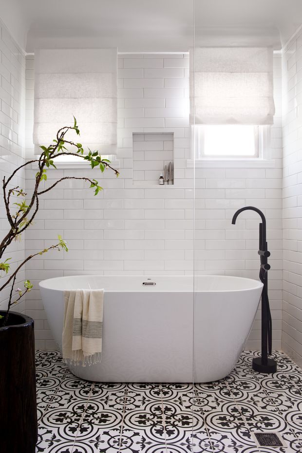 Bright, compact bathroom featuring a freestanding white bathtub, white subway-tiled walls, patterned black-and-white floor tiles, a black floor-mounted bath filler, roller blinds, and a potted branch adding an organic accent.