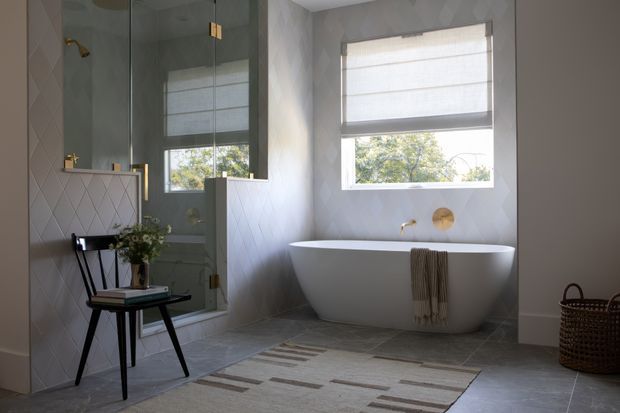 A refined bathroom featuring a freestanding white bathtub beneath a window with a roller blind, a glass-enclosed shower, and a wall-mounted basin with brass fittings. Light geometric wall tiles, grey floor tiles, a patterned neutral rug, and warm timber and woven accents create a calm, contemporary interior.