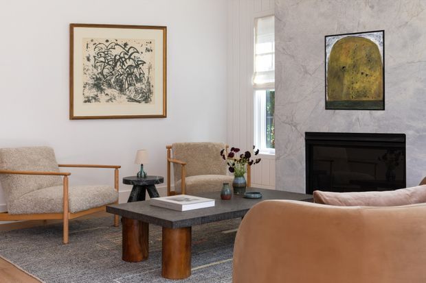 A refined living room with a large marble-look feature wall and black fireplace, neutral upholstered timber-framed armchairs, a caramel leather sofa, and a dark stone coffee table with chunky timber supports. Artwork, a narrow window, rug, and small decorative accents complete the calm, contemporary setting.