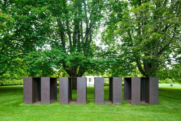 An outdoor architectural installation composed of several tall rectangular corten-steel-like sculptural panels arranged across a manicured lawn beneath mature leafy trees. The dark rust-brown metal forms create deep recessed openings and frame views toward a pale building in the background. The setting is a landscaped garden or estate grounds with a calm, contemporary sculptural character.