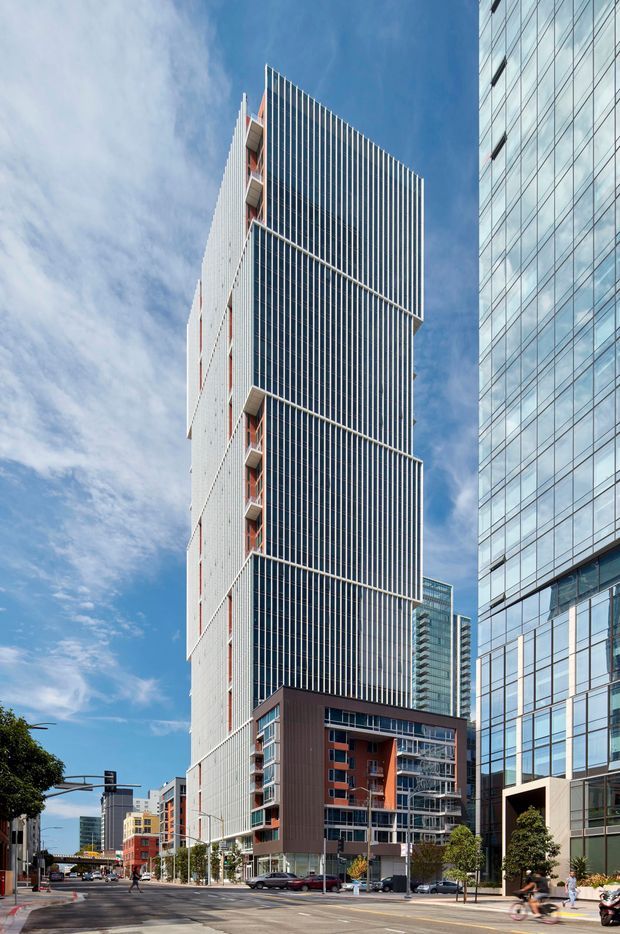 Street-level view of a contemporary high-density urban precinct, dominated by a tall narrow tower with a white facade and extensive dark vertical fins over glazed elevations. A lower multi-storey podium or residential building with dark brown cladding and red-orange balcony accents sits at its base, while a neighboring glass office tower frames the right side. Wide paved streets, traffic signals, street trees, and additional high-rise buildings establish a central-city commercial or mixed-use setting under a bright blue sky.