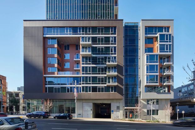 Street-level exterior photograph of a contemporary multi-storey mixed-use or apartment building in an urban setting. The facade combines extensive blue-tinted glazing, dark metal panels, warm reddish-brown wall sections, projecting balconies, and strong rectangular volumes. A taller glazed tower rises behind the main frontage, with a recessed vehicle entry at ground level and commercial or lobby glazing along the street.