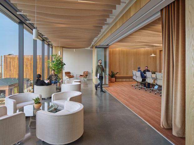 A spacious contemporary commercial office interior with an open lounge and collaborative meeting areas. Curved upholstered lounge chairs and small round tables sit beside full-height glazing, while a glazed meeting room with a long table and office chairs occupies the wood-lined area to the right. The space features warm timber ceilings and walls, dark polished flooring, slim dark window frames, pendant lighting, and floor-to-ceiling curtains.