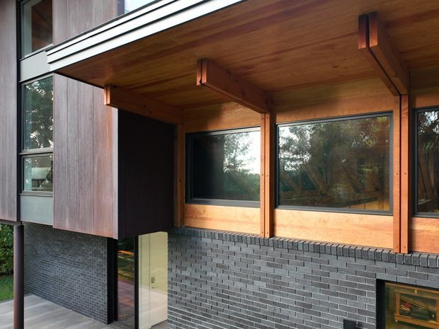 Close-up exterior view of a contemporary multi-level building corner, featuring dark grey brickwork at the lower facade, vertical dark timber cladding, warm timber soffits and exposed timber structural beams, and large dark-framed windows. A partially visible glazed entry door and recessed lower-level opening are present. The image emphasizes the facade materials and projecting covered structure.