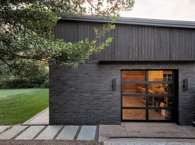 Exterior view of a contemporary single-storey building with a dark charcoal brick lower facade, vertically clad dark timber upper wall, and a black-framed glazed entry door. Concrete stepping slabs and a linear drain form the approach, with established greenery and lawn beside the structure. Warmly lit interior spaces are visible through the glazing.