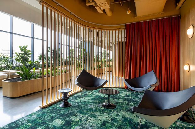 A stylish contemporary lounge or reception area with floor-to-ceiling glazing, abundant indoor planting, sculptural black lounge chairs, small round tables, and a richly patterned green rug. A curved timber slat screen partially separates the seating zone from the bright window edge, while dramatic red curtains and warm wood-toned walls create a strong feature backdrop. Exposed services and ductwork contribute an industrial-influenced commercial interior character.