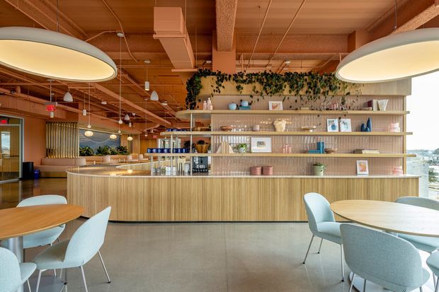 A bright contemporary hospitality or workplace café/lounge with a long timber-fronted service counter and open shelving displaying cups, bottles, books and plants. Pale upholstered dining chairs and round timber tables occupy the foreground. The space has a warm timber ceiling with exposed services and large oval pendant lights, while greenery and decorative objects soften the shelving. Glazing at the right provides an urban outlook.