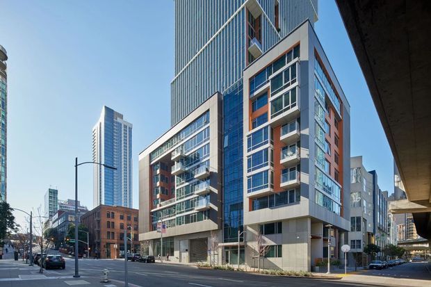 Street-level view of a contemporary multi-storey urban apartment or mixed-use building, with a tall blue-glazed tower behind a lower podium. The main facade combines extensive curtain-wall glazing, projecting balconies, pale metal or concrete panels, and reddish-brown accent panels. Dense city streetscape, adjacent high-rises, and an elevated roadway frame the scene.