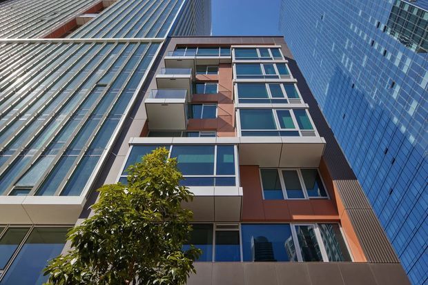 Upward-looking view of a contemporary multi-storey urban building framed by tall glass-and-steel towers. The central facade combines large blue-tinted windows, projecting balconies, and reddish-brown and grey panels, with a street tree partially obscuring the lower levels.