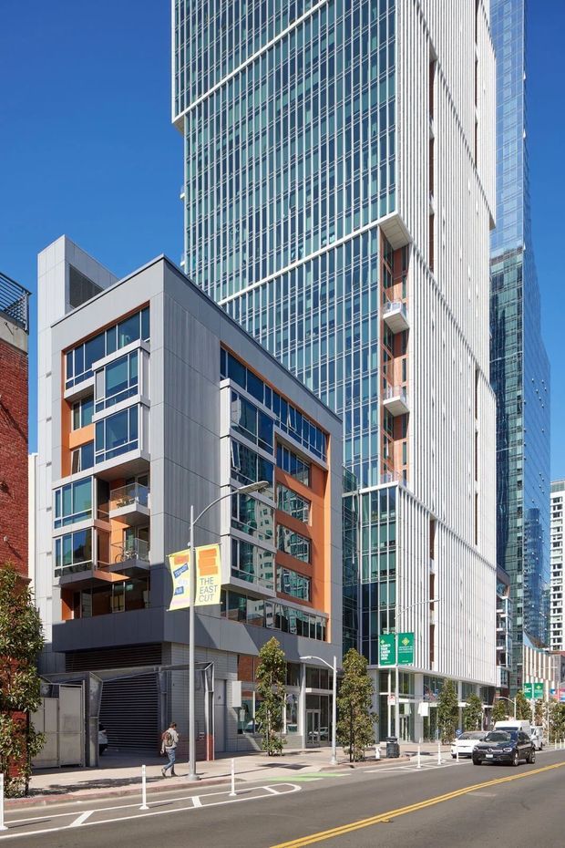 Street-level exterior photograph of a dense modern urban development, showing a mid-rise apartment or mixed-use building in the foreground beside a much taller glass-clad tower. The foreground facade combines grey metal panel cladding, extensive glazing, orange-brown accent panels, balconies, and ground-level entrances. The streetscape includes contemporary paving, bollards, street signage, and adjacent high-rise buildings under a clear blue sky.