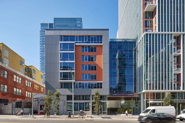 Street-level view of a dense urban commercial and mixed-use precinct, featuring several contemporary mid- and high-rise buildings with glass curtain walls, vertical fins, colourful panelled facades, recessed balconies, and active ground-floor retail or lobby spaces. Vehicles and pedestrians occupy the foreground streetscape.