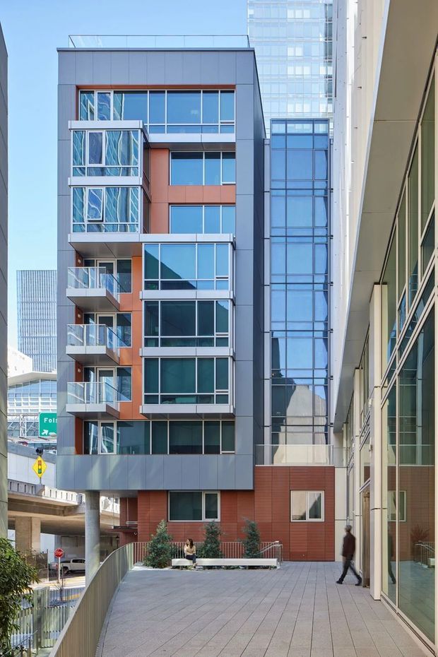 A modern multi-storey urban apartment or mixed-use building viewed from a raised pedestrian terrace. The facade combines grey metal panel cladding, reddish-brown panels, extensive blue-tinted glazing, projecting glass-railed balconies, and a flat roofline. A glass-enclosed vertical circulation or connecting structure is visible behind the main building, with neighbouring commercial architecture and an elevated roadway in the background.