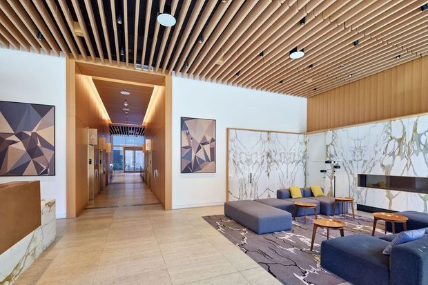 A spacious contemporary lobby or living area with a long timber-lined corridor, open-plan seating zone, large-format pale stone flooring, dramatic veined marble wall panels, and a linear fireplace or feature recess. The ceiling features an extensive exposed timber batten screen with integrated downlights and dark acoustic backing. Upholstered lounge seating, small timber tables, abstract artwork, and a patterned rug create a refined hospitality-like interior.