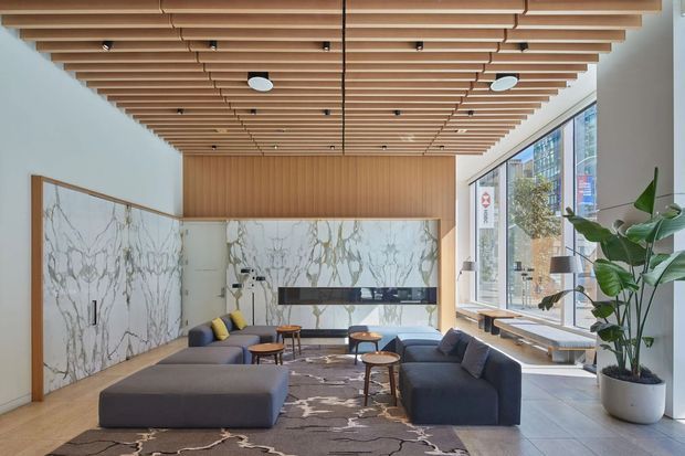 A bright contemporary lounge or reception area with a low modular sofa arrangement, timber flooring, patterned area rug, round coffee tables, and extensive floor-to-ceiling glazing. The space features a dramatic veined marble wall treatment, warm timber slatted ceiling with integrated lighting, timber wall panelling, and large indoor plants. Urban buildings are visible through the windows.