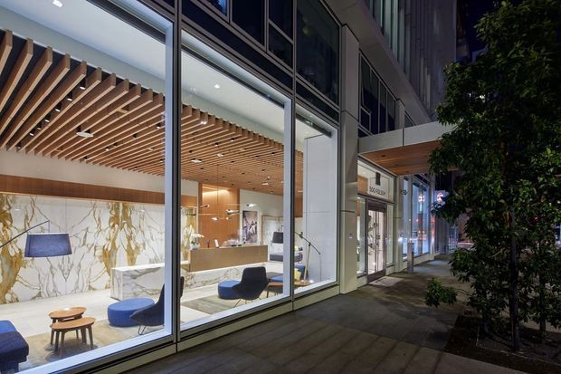 Street-level exterior view through full-height glazing of a contemporary commercial lobby or reception space. The interior features a warm timber slatted ceiling, dramatic white veined stone wall cladding, a reception desk, lounge seating, and pendant lighting. The building facade combines extensive glass with light-coloured structural framing, while a covered entrance canopy and planted streetscape are visible outside.