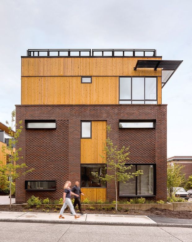 Street-facing view of a narrow multi-storey contemporary townhouse with a strong stacked facade composition. The lower level is clad in dark red-brown textured brick, while the upper level is clad in vertical warm timber boards. Large black-framed windows, a timber entry door, rooftop railing, and a projecting upper-level canopy are visible. The building sits on a landscaped urban streetscape with a sidewalk and small planted trees.