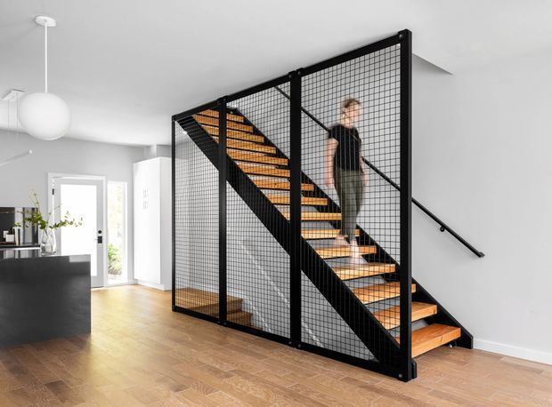 Bright contemporary open-plan interior featuring a prominent black metal staircase with timber treads and fine wire-mesh balustrade/enclosure. The stair rises through the centre of the room, with a person ascending it. A black kitchen island and partial kitchen are visible to the left, along with white walls, light timber flooring, minimalist spherical pendant lighting, and a glazed door. The image is primarily focused on the staircase and circulation space.