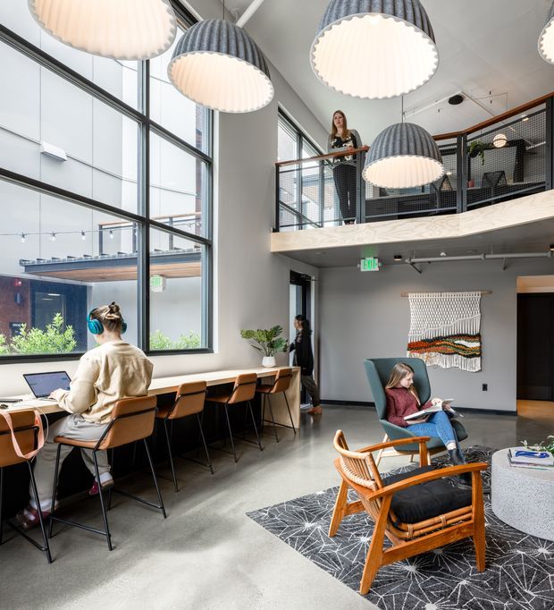 Bright contemporary commercial lounge and workspace with a double-height atrium, mezzanine level, expansive industrial-framed glazing, concrete-look flooring, and oversized ribbed pendant lights. A long communal work counter runs along the windows, while lounge chairs and a patterned rug create a relaxed seating area. Exposed black mesh mezzanine balustrading, timber handrail, woven wall art, and indoor planting add warmth to the otherwise modern shared interior.
